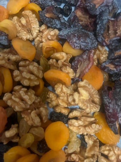 This is a close-up, overhead shot of a mixture of dried fruits and nuts. The subjects include dark, wrinkled dried plums, bright orange dried apricots, light brown walnuts, and some smaller, lighter colored raisins. The background appears to be a light blue surface, possibly fabric. There are no people or actions depicted, and the image is a still life of food items. There is no information about Edem, Russia, or any urban environment.
