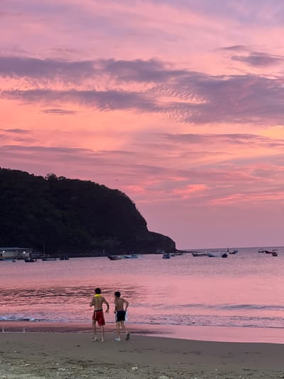 The image depicts a serene sunset scene at the beach in San Juan del Sur, Nicaragua. Two young males are walking along the sandy shore, facing away from the camera. They are wearing swim trunks, and one has a yellow band around his neck. The ocean is calm with gentle waves lapping at the shore, reflecting the vibrant pink and purple hues of the sky. In the distance, a tree-covered hill juts out into the water, casting a dark silhouette against the colorful sky. Several small boats are anchored in the bay, adding to the tranquil atmosphere. The sky is filled with soft, wispy clouds painted in shades of pink, lavender, and orange, characteristic of a tropical sunset. The overall impression is one of peacefulness and natural beauty.