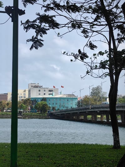 The image displays a daytime urban riverine scene in Thuận Hóa Ward, Hue, Vietnam, under a partly cloudy sky. The foreground consists of green grass. A vertical dark green pole with a dark fixture is prominent on the left. Silhouetted dark tree branches with sparse foliage are visible against the sky from trees in the immediate foreground, on both the left and right.
A body of blue-green water with light ripples occupies the midground. A multi-span concrete bridge traverses the water from left-center to right, featuring light-colored railings and numerous streetlights. Several indistinct human figures are discernible on the bridge, along with at least one light-colored vehicle, appearing to travel towards the right.
On the far bank, a prominent light blue, multi-story building with multiple windows is situated behind the bridge. To its left and further back, a taller, light grey multi-story building is visible. On the roof of this grey building, a dark sign with white lettering clearly reads "BỆNH VIỆN TRUNG ƯƠNG HUẾ" (Hue Central Hospital). Two red Vietnamese flags, each with a central yellow star, are flying from poles on a rooftop located to the right of the grey building. Additional buildings and scattered green foliage line the distant riverbank.