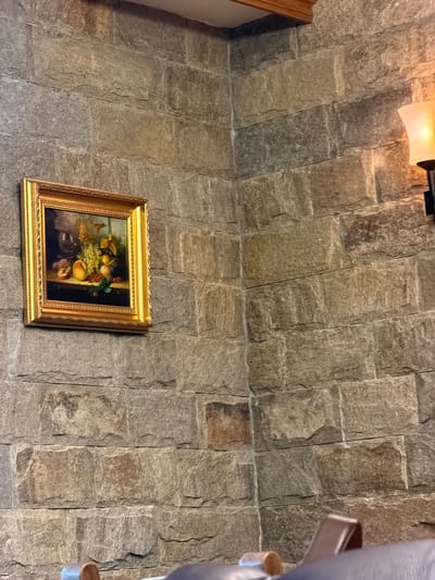 A framed still life painting hangs on a rough stone wall. The painting has a gold frame. The wall appears to be part of an interior. A light fixture is visible near the top of the image. Part of a chair or other furniture is visible on the right side. The location is likely in Кирково, Bulgaria.