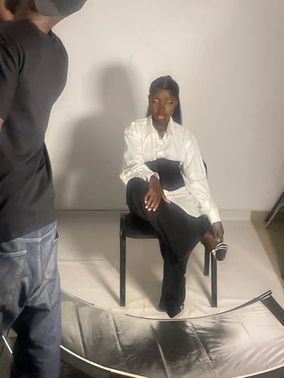 In a studio setting with a plain white backdrop, a woman with dark skin, wearing a white collared shirt, a black corset, and black wide-leg pants, sits on a black chair. Her hair is pulled back into a ponytail, and she has bold eye makeup. One leg is crossed over the other, and she wears open-toed sandals. To the left of the frame, a person wearing a black t-shirt and blue jeans is partially visible, facing away from the camera. The floor is covered with a reflective surface, likely a photo backdrop or floor mat. The scene appears to be a professional photoshoot. The image does not contain information that allows for the precise determination of its location within the city of South Ropp, Nigeria, or any other specific city.