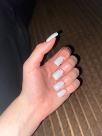 A hand with square-shaped, white acrylic nails rests on a brown, ribbed surface. The hand is positioned in the foreground of the image, with the fingers slightly curled. The background is dark and out of focus, with a hint of black fabric to the left and the ribbed surface continuing to the right. The scene is dimly lit, with a soft glow illuminating the nails. The location is not specified in the image.