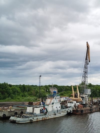A cloudy day at the Vytegra River port, Russia. Two barges are moored side-by-side, their weathered decks laden with industrial equipment and what appears to be construction materials. A large, towering crane dominates the scene, its metallic arm reaching towards the overcast sky. Next to it, a smaller vessel, possibly a tugboat, is docked, bearing the markings "PT-329" on its hull. In the background, a dense line of green trees marks the edge of the land, with a few distant buildings and a telecommunications tower visible. The water in the foreground is calm, reflecting the muted tones of the sky and the surrounding industrial landscape.