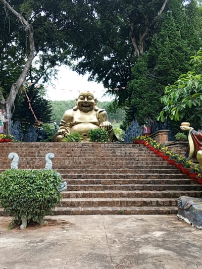 The image shows a large golden Buddha statue seated at the top of a wide stone staircase in Nam Ban, Vietnam. The Buddha statue is smiling and appears to be made of a reflective material. The staircase is lined with red planters filled with yellow flowers on the right side. To the left of the staircase, there is a hedge sculpture with two stone elephant-like figures. In the background, behind the Buddha, are more statues and lush green trees, with a hillside visible in the distance. Prayer flags are strung across the trees. The ground in front of the staircase is paved with concrete.