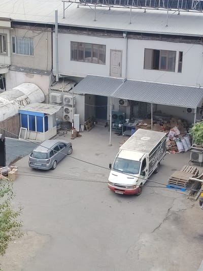The image depicts an outdoor industrial or commercial area in Khujand, Tajikistan. Two vehicles are present: a silver station wagon parked on the left and a white van with red trim parked on the right, facing left. The station wagon has a license plate that is partially obscured. The van has text and graphics on its side.
The background features a multi-story building with a light-colored facade and a metal roof. Several windows are visible on the building's upper floor. Below the upper floor, there is a covered outdoor area with a metal roof supported by pillars. Air conditioning units are mounted on the building's exterior.
To the right of the van, a stack of wooden pallets is visible. In front of the covered area, there are piles of cardboard boxes and bags, suggesting a storage or loading area. To the left of the station wagon, there is a small blue and white structure with a metal roof, and some discarded window frames are leaning against it.
The ground surface is paved with asphalt, showing signs of wear and cracking. Overhead power lines or cables are strung across the scene. Green foliage from a tree is visible in the lower left corner of the image. The overall impression is that of a functional, somewhat unkempt, working environment.