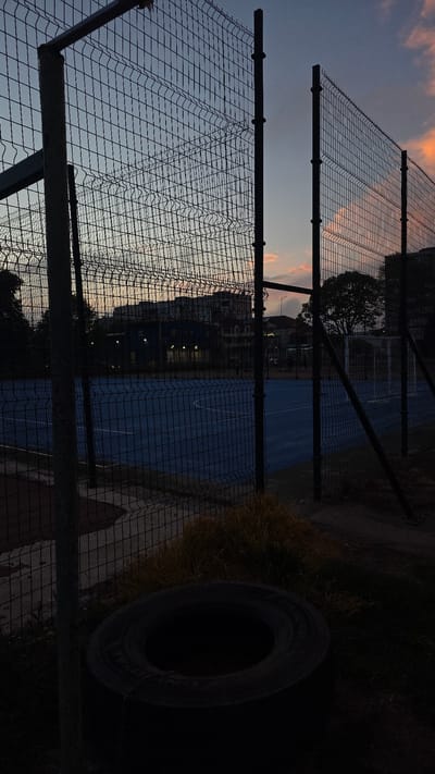 The image shows a fenced tennis court in Kavarna, Bulgaria at dusk. The fence is made of chain-link, with metal posts. A tire is visible to the right of the fence, surrounded by overgrown grass and vegetation. In the background, there are buildings and trees silhouetted against the twilight sky, which has hues of orange and blue. The tennis court itself is blue and appears to be empty.