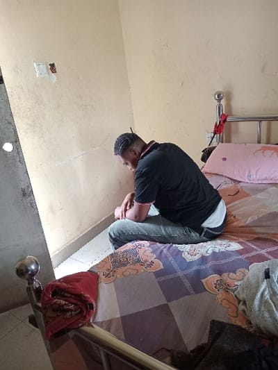 An individual is depicted seated on the edge of a bed within an indoor setting in Umueshi, Nigeria. The person, appearing to be a young adult male with braided hair, wears a black collared shirt with red and white trim, blue jeans, and a white undershirt. He is positioned with his head bowed and hands clasped near his knees, facing towards the left of the frame. The bed features a polished metal frame with spherical finials and a mattress covered by a patterned bedspread displaying geometric squares in brown, gray, and purple, interspersed with orange and beige ornamental designs. A pink pillow with a light-colored animal silhouette is at the head of the bed, on which a dark object rests. A red patterned fabric is folded on the metal footboard on the left, and a crumpled light-colored fabric is on the bed's right side. The room's walls are painted pale yellow/beige, exhibiting discoloration and dirt marks. An electrical switch plate with a broken or missing switch and exposed wiring is visible on the left wall. A red ribbon or cloth is tied to the right headboard finial. The floor, partially visible on the left, appears to be light-colored tile.
