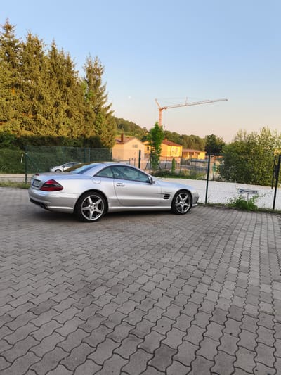 A silver Mercedes-Benz SL-Class roadster is parked on a paved lot. The car is viewed from the rear three-quarter angle. The lot is made of grey interlocking pavers. Behind the car is a chain-link fence with a white mesh cover. Beyond the fence are trees, buildings, and a construction crane in the distance. The sky is clear and blue, with a faint crescent moon visible. The sun is low in the sky, casting long shadows, suggesting it is either early morning or late afternoon. A sign with "P Reserviert" is visible on the fence, indicating a reserved parking spot. The scene appears to be in Pegnitz, Germany. There are no people visible in the image.