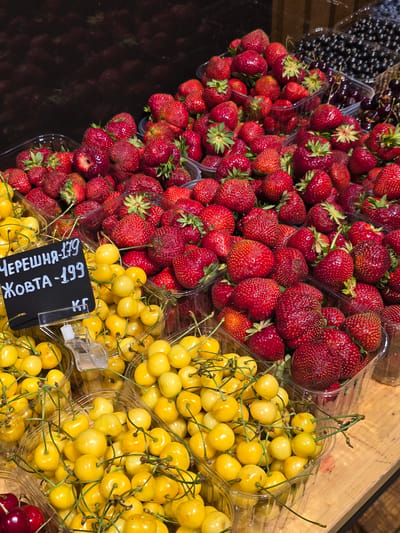 The image displays a vibrant overhead view of fresh fruit, primarily strawberries and yellow cherries, arranged in clear plastic containers. The strawberries are a rich red, some with visible green stems and leaves, filling several containers in the foreground and mid-ground. To the right and slightly further back, piles of bright yellow cherries are stacked in similar containers, their stems still attached. In the upper left corner, a container of dark, small berries, likely blueberries or currants, is partially visible. The fruits are laid out on what appears to be a wooden surface, suggesting a market stall or a retail display.
A small black price tag is affixed to one of the containers of yellow cherries, featuring white handwritten text in Cyrillic. The visible text reads "ЧЕРЕШНЯ - 179" and "ЖОВТА - 199 КГ". This translates to "Cherry - 179" and "Yellow - 199 KG". The lighting suggests it could be daytime, with the light casting slight shadows, indicating an indoor or shaded outdoor setting. There are no people visible in the frame. The overall impression is one of fresh produce ready for sale, likely at a farmer's market or grocery store in Petropavlivska Borshchahivka, Ukraine. The scene evokes a sense of freshness and abundance.