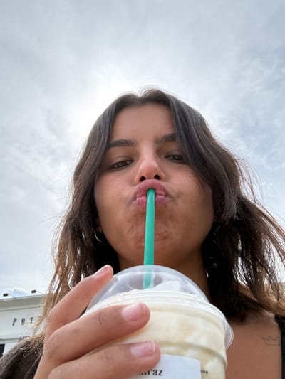 An outdoor, low-angle image depicts a young adult female from the chest up, positioned against an overcast sky. The subject has dark hair, dark eyes, and a light-medium complexion. Her lips are pursed around a teal plastic straw, actively consuming a light-colored, blended beverage from a clear plastic cup with a domed lid, consistent with a Starbucks Frappuccino. Her right hand grips the cup, with fingers visible. A small outline tattoo of a crown is visible on her left collarbone/shoulder area. To the lower left, in the background, a white building with dark lettering "POTTERY BARN" is partially visible. This scene is located in West Windsor, United States, specifically near the Pottery Barn at Nassau Park Pavilion.