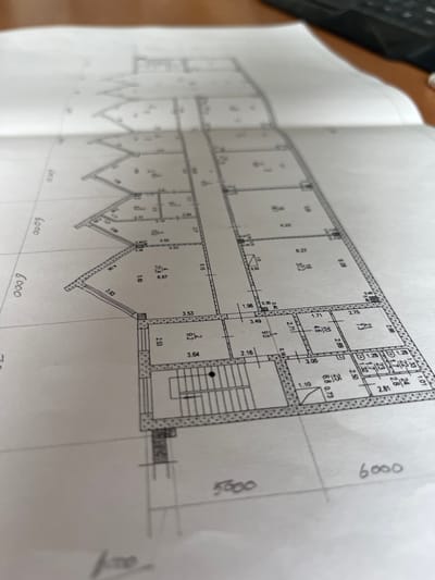 The image shows a close-up, angled view of architectural blueprints spread open on a surface, possibly a desk. The blueprints depict a floor plan with various rooms, corridors, and staircases, indicated by lines and labels. Numerical values, likely representing dimensions or areas, are present within the rooms. Faint grid lines are visible, suggesting a scaled drawing. A portion of a dark keyboard is visible in the upper right corner, and the background appears to be a wooden surface. The image is in focus on the blueprints. There is no visible indication of the city of Aktau, Kazakhstan, or any people.