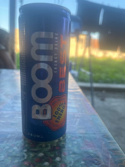 A close-up, eye-level, outdoor shot features a blue aluminum can of "BOOM BEST ENERGY DRINK" placed on a table with a patterned tablecloth. The can is oriented vertically and takes up most of the left side of the frame. Its label has large silver lettering spelling out "BOOM" and "BEST" in orange. To the right of the word "BEST" is text indicating "ENERGY DRINK." Below this, an orange circular logo reads "BEST QUALITY 100% ENERGY" with smaller text in an orange banner underneath that says "BEST TASTE." The can is slightly obscured by a bright, hazy light coming from the right, which also blurs the background. The background suggests an outdoor environment with greenery and structures, possibly a yard or park, but it is too indistinct to identify specific features or definitively place it in Jrarbi, Armenia. The table surface has a reflective quality and displays a colorful, abstract pattern with red and green elements. The lighting is bright and natural, suggesting daytime. No people are clearly visible in the image.