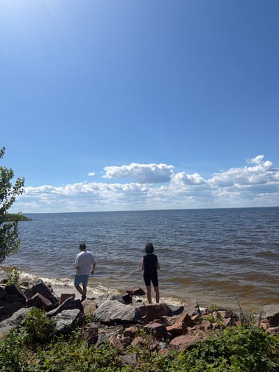 The image shows two young men standing on a rocky shore next to a body of water. The scene is set in a natural environment, likely a lake or river, under a bright blue sky with fluffy white clouds. The sun is shining, suggesting it is daytime. The man in the foreground is wearing a white t-shirt and light-colored shorts, while the man further back is in a black t-shirt and dark shorts. They appear to be casually enjoying the outdoors. The water is rippling, indicating a gentle breeze. The rocky shore is covered in green vegetation, adding to the natural feel of the location. There are no visible objects or text in the image.