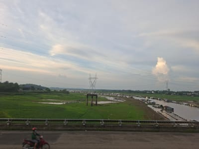 This image captures a wide-angle view of a rural landscape in Dong Nai City, Vietnam, likely during daylight with overcast skies. The foreground features a concrete bridge with a metal railing, and a blurred figure on a red motorcycle wearing a green shirt and red helmet is seen riding on the road below the bridge.
The middle ground depicts a vast expanse of green grassland with scattered areas of shallow water, suggesting a recently flooded or marshy terrain. A prominent pylon with thick power lines stretches across the scene, contributing to the industrial infrastructure. Further into the distance, a river or canal is visible, lined with numerous stilted houses and rafts, indicating a community living on the water. The houses are of various sizes and construction, with some appearing to be floating or built on platforms.
The background shows a treeline bordering the river and rolling hills in the distance. The sky is a muted grey with wisps of clouds, casting a soft, diffused light over the entire landscape. The overall impression is one of a natural environment interwoven with human habitation and industrial elements, characteristic of many rural areas in Southeast Asia. The presence of the power lines and the stilted houses suggests a developing area where traditional ways of life coexist with modern infrastructure.