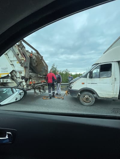 The image depicts the aftermath of a collision between a white van and a garbage truck on what appears to be a road in or near Korabsel'ki, Russia. Two individuals, possibly involved in the accident or providing assistance, are standing between the two vehicles. One is wearing a red jacket and the other is wearing a gray jacket. The front of the white van shows visible damage, and the rear of the garbage truck is also damaged. The sky is overcast. The photo appears to be taken from inside another vehicle.