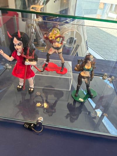 The image shows a display case with three collectible figures. On the left, a doll with black hair, red devil horns, and a red dress is holding a trident. In the middle, a blonde figure in a striped top, shorts, and a hat is posed with claws. On the right, a figure in a brown jacket, green top, and ripped pants wears a hockey mask and holds a weapon. A price tag of "$120.99" is visible on the trident holder, and another of "$180.00" is on the base of the hockey-masked figure. A black combination lock is present on the dark blue surface of the display. The background reveals a blurry scene of what appears to be a retail environment with large statues or displays.