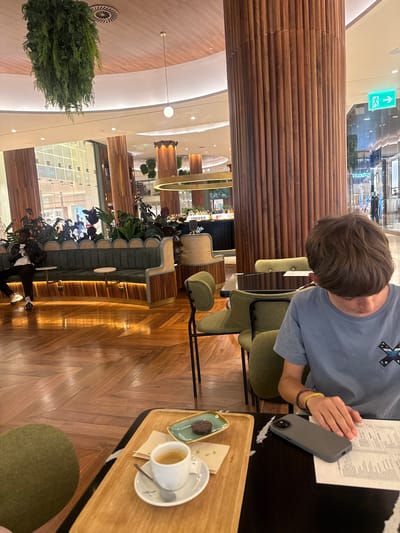 The image captures an indoor scene within a modern establishment, likely a cafe or restaurant, in Matosinhos, Portugal. The ambiance is sophisticated, with polished wooden floors laid in a herringbone pattern and large, vertically ribbed wooden columns dominating the space. A lush, hanging plant arrangement drapes from the ceiling, adding a touch of nature to the interior.
In the foreground, a young person wearing a light blue t-shirt is seated at a table. They are looking down at a menu and have a smartphone with a gray case resting on the table. A small white cup containing a dark liquid, presumably coffee, sits on a saucer beside a piece of dark chocolate on a small green dish, all placed on a wooden tray. Another person, dressed in a black leather jacket and white t-shirt, is seated on a long, dark green banquette in the middle ground. This banquette is set against a backdrop of potted plants and features a woven cane accent on its sides.
The background reveals a retail environment, with storefronts visible through large glass windows, suggesting the establishment is situated within a shopping mall or a similar commercial complex. An illuminated green exit sign with an arrow is mounted on one of the wooden columns. The lighting appears to be a mix of ambient and focused light sources, creating a warm and inviting atmosphere. The scene depicts a moment of leisure, with individuals enjoying refreshments and the surroundings.