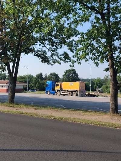 In Hluboká nad Vltavou, Czechia, on a clear day, two trucks are parked in an open lot. The truck on the left is blue, and the truck on the right is yellow with a large tank on its back that has "SAT" written on it. A building with a red roof and white walls, labeled "WC," is visible in the background. Trees with green leaves surround the area, and a paved road is in the foreground. The scene is calm and depicts a typical roadside rest area.