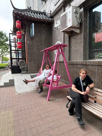 In Hunchun, China, on a cloudy day, a woman in a white dress and camouflage pants sits on a pink swing, her legs outstretched. Next to her, a child wearing a white shirt is also on the swing. To the right of the swing, another woman wearing a black t-shirt and black pants sits on a wooden bench, holding a purse. The scene takes place outdoors, on a sidewalk paved with interlocking bricks. In the background, a building with traditional Chinese architectural elements, including a curved roof and red lanterns, is visible. An air conditioning unit is mounted on the building's exterior.