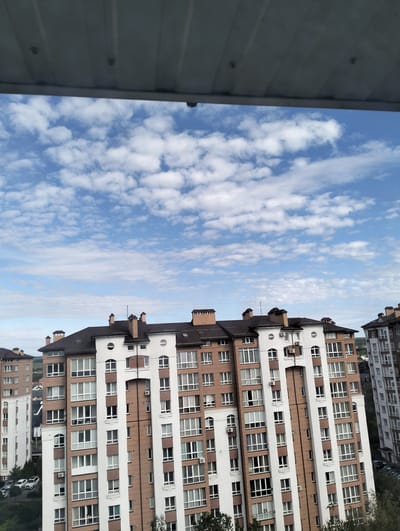 The image shows a view of multi-story apartment buildings in Krykhivtsi, Ukraine, taken from an elevated perspective, possibly a balcony or higher floor of another building. The sky is predominantly blue with scattered white clouds. The buildings are constructed with a combination of white and brown exterior finishes and have numerous windows and rooftop chimneys. Vegetation and parked cars are visible at ground level. A portion of a ceiling or roof overhang is visible at the top of the frame, indicating the vantage point of the photograph. No people are directly visible in the frame.