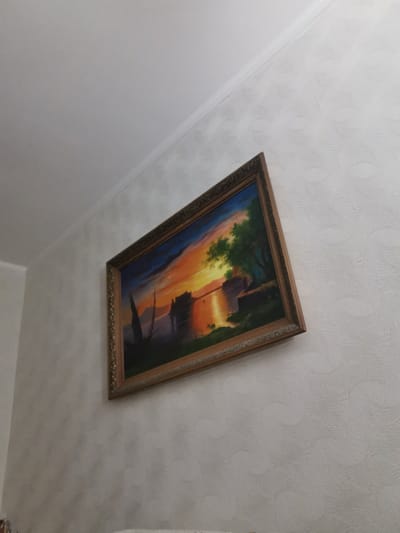 A framed painting of a sunset over a body of water is displayed on a white textured wall. The painting depicts a vibrant sky with hues of orange, yellow, and blue, reflecting on the water. Silhouetted trees and what appears to be structures or boats are visible on the water's edge. The painting is hung at an angle, with the top of the frame closer to the ceiling than the bottom. The wall has a repeating pattern of curved lines. The scene appears to be indoors, possibly in a residential setting in Pociumbeni, Moldova.