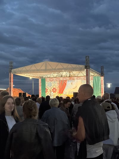 An outdoor event is taking place at dusk in the city of Uzda, Belarus, in front of a stage. The stage has a white roof supported by scaffolding, and the backdrop features a colorful, embroidered pattern with green fabric on either side. A performer in a white outfit is on stage, possibly singing or speaking, with two white birds depicted in flight above them on the backdrop. A banner with Cyrillic text, which reads "Са святам" (meaning "Happy Holiday"), is visible on the stage.
A crowd of people is gathered in front of the stage, facing towards it. They appear to be watching the performance and enjoying the event. The sky above is dark and cloudy, indicating either a late evening or approaching bad weather. In the background, there are buildings with red tiled roofs and streetlights. The overall scene depicts a community gathering and celebration in the city of Uzda.