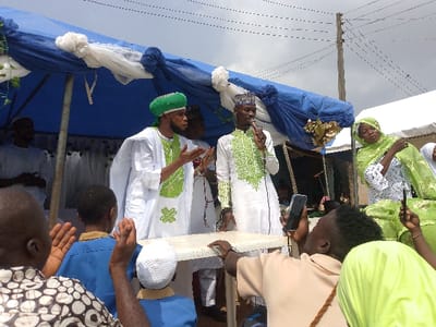 An outdoor gathering is depicted in Ijoko, Nigeria, beneath a blue and white fabric canopy. In the foreground, two men in white traditional Islamic attire are positioned. The man on the right, wearing a white robe with green embroidery and a patterned cap, holds a microphone to his mouth, speaking. The man to his left, wearing a similar white robe with green embroidery and a green turban, gestures with open hands. A white plastic table is situated in front of them. In the midground, a woman in a green hijab and white embroidered top is visible to the right.
The audience consists of multiple individuals, with their backs or partial profiles visible in the lower portion of the frame. Several audience members are raising their hands, while others are using mobile phones to record the event. Notably, a person in a beige shirt in the lower right foreground holds a smartphone pointed upwards. Overhead, utility wires crisscross against a bright sky, with a wooden utility pole visible in the upper right background. The setting appears to be a public or community event under temporary shelters.