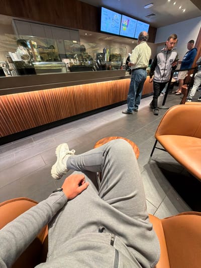 The image shows the interior of a Starbucks in North Bethesda, Maryland, USA. A person is seated in a tan leather chair, wearing gray sweatpants, a gray sweatshirt, and white sneakers. They are resting their arm on their leg. In the background, other customers are ordering at the counter. A backlit menu board is visible above the counter, and various beverage preparation equipment can be seen through the glass. The decor includes warm-toned wood paneling.