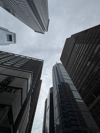 The image shows a low-angle perspective of several skyscrapers in Philadelphia, United States. The sky is overcast. There are five main buildings in view, each with a distinct architectural style. The tallest building on the right has a stepped facade with reflective glass panels. The building in the bottom right has a large sign on its lower floors. Another building is visible in the bottom left, with a distinctive rectangular opening near its top. A tall, slender building with vertical stripes appears on the left. At the top center is a building with a grid-like window pattern, giving a darker appearance.