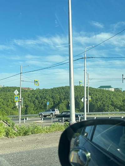 This is a medium shot, captured from a vehicle, looking out towards a tree-covered hill on a sunny day in Svetly, Russia. The sky is bright blue with wispy clouds and a tall, white lamppost dominates the left side of the frame. To the right of the lamppost, there are electrical wires strung across the scene.
Further into the midground, a silver SUV is parked on the side of a road, with a black sedan positioned behind it. Both vehicles appear to be stationary. There are yellow pedestrian crossing signs with a white walking figure on them visible on poles near the road. A horizontal metal railing runs along the edge of the road, separating it from a patch of green vegetation. In the background, a large building with a green roof is visible atop the hill, partially obscured by the dense foliage of trees. The foreground shows a portion of the road and the side mirror of the vehicle from which the image is taken. The mirror reflects the blue sky and some of the vehicle's interior.