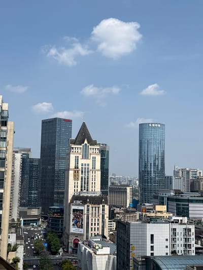 The image shows a cityscape in Jinjiang District, Chengdu, China. Several high-rise buildings are visible. One tall, reflective glass tower features Chinese characters, likely a building name. A second prominent building is identified as the JW Marriott. Adjacent to the JW Marriott is a building with a pointed roof structure. A KBC billboard is visible on one of the buildings. Trees line streets below. The sky is light blue with scattered white clouds. Cars move along the streets.