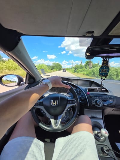 The image is a first-person perspective from inside a car, looking forward and slightly to the left. The driver's hands are on the steering wheel, and they are wearing a white watch. The car's interior is visible, including the dashboard with a Honda emblem on the steering wheel, a gear shift, and a car air freshener hanging from the rearview mirror. Outside the car, the road stretches ahead, flanked by green vegetation under a bright blue sky with scattered white clouds. Another car is visible in the distance on the road. The location is identified as Dalgopol, Bulgaria.