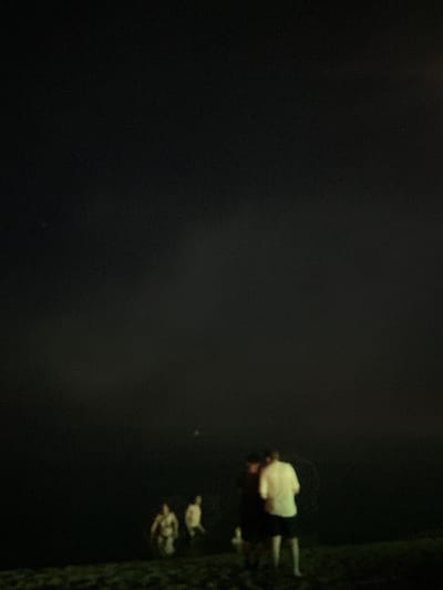 The image depicts a dimly lit, possibly nighttime scene with several blurry figures in the foreground, set against a dark, hazy background. In the lower right quadrant, two individuals are visible. One, facing away from the camera, wears a white shirt and dark shorts. To their left and slightly behind, another figure is partially visible, also appearing to wear dark clothing. Further to the left, two more figures are present, one wearing a light-colored garment and the other a darker one. The ground in the foreground appears to be covered in grass or vegetation. A faint light source is discernible in the upper right portion of the frame, suggesting artificial lighting or possibly the moon. The overall impression is one of a clandestine or casual gathering in an outdoor, low-light environment. The location is identified as Canet de Mar, Spain, which is a coastal town. This context suggests the scene might be taking place near the beach or a waterfront area.