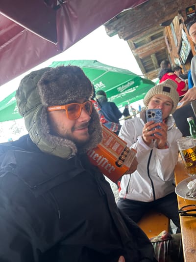 The scene depicts an outdoor or semi-enclosed seating area at what appears to be a ski resort establishment in St. Christoph, Austria.
In the foreground, a male subject is seated at a light-colored wooden table. He wears a dark olive green ushanka-style hat with brown fur trim, orange-tinted sunglasses displaying "Jägermeister ORANGE" on the frame, and a black winter jacket. He has a beard and is looking towards the right side of the frame with a slight smile. Under his left arm, he holds a six-pack carrier of "Jägermeister ORANGE" mini bottles or cans.
To his right, a female subject is also seated at the wooden table. She wears a light-colored knitted beanie and a white winter jacket. She holds a light blue patterned smartphone with both hands, aiming it towards the male subject, consistent with taking a photograph or video.
On the table to the female's right, a large glass of beer with "Hofbräu" visible on its side, a small green glass bottle, a white napkin or food item on a plate, and a pair of black eyeglasses are present.
The background shows several blurred individuals in winter attire, including one in a red jacket and another in a blue jacket. Overhead, a dark red tarpaulin or awning provides cover, and green umbrellas bearing "Mönchhof" and other German text are visible further back. A rustic wooden building structure is partially visible on the upper right, with signs affixed to it. The sky appears overcast, suggesting diffused natural light.