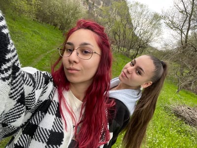 Two women pose outdoors. The woman on the left has long brown hair and wears a grey hooded sweatshirt. The woman on the right has red hair and wears eyeglasses and a black and white checkered sweater. They are standing in a grassy area with trees in the background. The location appears to be Barkach, Bulgaria.