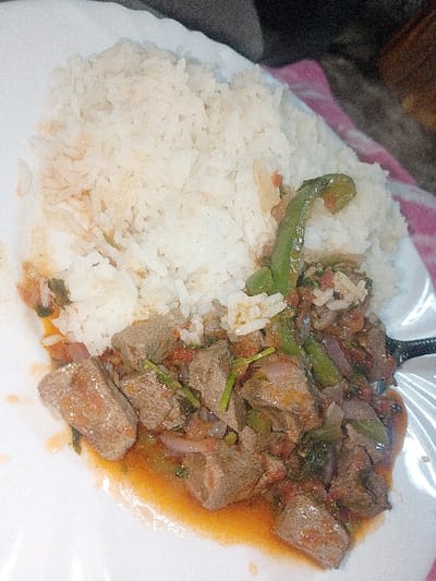 The image displays a close-up overhead view of a meal served on a white plate, possibly prepared or consumed in Limuru Central ward, Kenya. The plate is divided, with a heap of white, cooked rice occupying the upper left and central portions. Adjacent to the rice, filling the lower right, is a stew containing numerous brownish-grey, irregularly shaped chunks of what appears to be cooked organ meat, likely liver or tripe. This stew is mixed with various chopped vegetables, including diced red onions, diced red tomatoes, and several elongated green vegetables resembling green beans or long chili peppers. Green herbs are also visible throughout the stew, which is coated in a glossy, orange-red sauce, with some liquid pooled at the bottom of the plate. A dark utensil handle is partially visible on the right edge of the stew. The white plate rests on a surface with a blurred pink and white checkered pattern. The background beyond the plate and immediate surface is dark and indistinct. No individuals are present, and no actions or interactions are depicted.