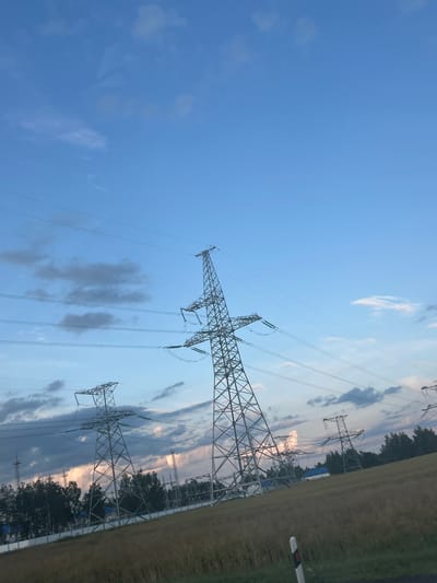 The image depicts a wide shot of a rural landscape with several tall electricity pylons and power lines under a cloudy blue sky. The foreground features a gently sloping field of dry grass and a small white and red traffic marker. In the mid-ground, dense green trees line the horizon, with some buildings and additional electrical infrastructure visible behind them. The pylons are constructed of latticed metal, with multiple power lines strung between them, stretching across the frame. The sky is a mix of clear blue with scattered grey and white clouds. The scene suggests an area on the outskirts of a populated region, possibly a power substation or a major transmission line route. The exact location within Старасвержанскі сельскі Савет, Belarus, is not specified in the image itself, but the overall setting is consistent with a rural or semi-rural environment in that region. There are no people visible in the image.
