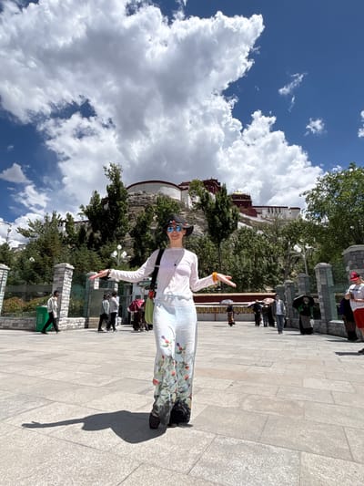 The image displays an outdoor scene on a bright, sunny day, featuring a woman posing in the foreground against the backdrop of a grand building complex. The image is oriented vertically but is presented on its side.
In the immediate foreground, a woman, likely the main subject, is captured mid-pose, smiling broadly at the camera with her arms outstretched. She wears a white long-sleeved top, light-colored pants or a long skirt with a vibrant floral and bird pattern, sunglasses, and a dark hat. A yellow bracelet adorns her right wrist, and she carries a shoulder bag. Her elongated shadow, clearly showing the outline of her arms, stretches across the paved ground to her right.
Behind her, majestically situated on a rocky hill covered with green trees and foliage, stands a large, multi-tiered building complex. This iconic structure, with its distinctive white walls, red sections, and smaller gold-colored roofs, is identifiable as the Potala Palace.
The ground in front of the palace is a spacious, light gray paved plaza made of stone tiles. Several other individuals, appearing to be tourists, are scattered across the mid-ground, some walking and others standing near stone pillars or low walls that delineate sections of the plaza. A green trash can is partially visible in the lower left background, bearing Chinese characters that translate to "Recyclable Waste" (可回收物).
The upper part of the image shows a clear blue sky dotted with large, fluffy white cumulus clouds, suggesting pleasant weather. The strong sunlight casts crisp shadows, confirming it is daytime. The overall scene depicts a lively tourist location in the Chengguan District, China, with the woman expressing visible cheerfulness.