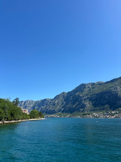 The image shows the landscape in Prčanj, Montenegro. On the right, there is a mountain range running along the coast, with a series of buildings and structures. The foreground features green vegetation and a shoreline. To the right of the shoreline is the body of water. To the left is a vast, clear blue sky.