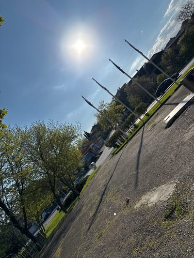 The image depicts a gravel parking area adjacent to a street and a line of flagpoles in Topoli, Bulgaria. The sky is bright blue with the sun shining. A tree is visible in the foreground with new leaves budding. Cars are parked along the street, and buildings can be seen in the background. A white object, possibly a ball, rests on the gravel. A red object is visible near the white ball. The flagpoles cast long shadows onto the gravel.