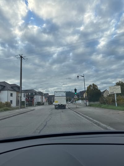 This image depicts a view from inside a vehicle on a roadway in Ammerschwihr, part of Kaysersberg Vignoble, France. The scene is observed under an overcast sky with patches of brighter cloud and limited visible blue sky.

The asphalt road, exhibiting faint lane markings, extends forward from the vehicle's dashboard. A white box truck, displaying "JH Transports" on its rear, occupies the right-center of the road ahead, moving away from the viewer.

To the left, a tall wooden utility pole with power lines stands beside the road. Beyond it, a row of multi-story houses with light-colored facades and dark gabled roofs are visible, interspersed with some greenery. A traffic light, showing red, is partially visible further down the road on the left.

To the right, a paved shoulder or sidewalk runs alongside the road. A black lamppost with a vintage-style fixture is present. Immediately past the lamppost, a traffic light displays a green signal. Further right, a large off-white sign advertises "Vins d'Alsace - Crémant DOMAINE STURN DÉGUSTATION - VENTE." Beyond the sign, vegetation and another house are visible, with distant green hills under the cloudy horizon. No individuals are visible.