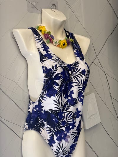 The image shows a white mannequin torso wearing a blue and white floral print one-piece swimsuit. A colorful crocheted flower necklace is draped around the neck of the mannequin. The mannequin is standing against a grey, textured wall with diagonal line markings. To the right of the mannequin, there is a white object with a relief of a bell on it. The scene appears to be indoors, possibly a retail display. The location of Ayn al Basha, Jordan is not evident from the image itself.