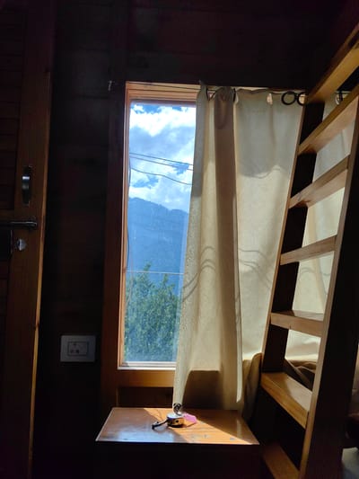 The image depicts an interior scene with a window offering a view of a mountainous landscape. The room appears to be rustic, with wooden walls and a wooden ladder leading to an upper level, likely a loft bed. A light-colored curtain with a subtle patterned texture hangs from a rod by the window. On a wooden surface in front of the window, a small, yellow lock with a key attached is visible. To the left of the window, there's a door with a metallic handle and latch, and a white electrical outlet on the wall. The overall lighting suggests it is daytime, with sunlight streaming through the window, casting shadows within the room. The exterior view shows lush green trees and a hazy mountain range under a cloudy sky with visible power lines. The image seems to be taken within a lodging in Siumsa, India, given the presence of the electrical outlet which is common in India.