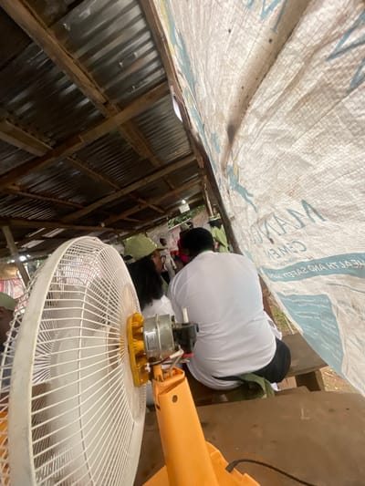 The image depicts an outdoor gathering under a makeshift shelter in Orun Ekiti, Nigeria. A large white oscillating fan with an orange base is positioned in the foreground, suggesting a need for cooling in the warm climate. Behind the fan, several individuals are seated on wooden benches. Most are wearing green caps and white shirts, possibly indicating a uniform or affiliation with an organization. One person in the center is facing away from the camera, wearing a white shirt and black pants. The shelter's roof appears to be made of corrugated metal supported by wooden beams. A large, white tarp with blue text, possibly advertising cement, is draped over part of the shelter, creating a semi-enclosed space. The ground beneath the shelter is visible and appears to be earthy. The overall impression is a casual, community-oriented event taking place outdoors.