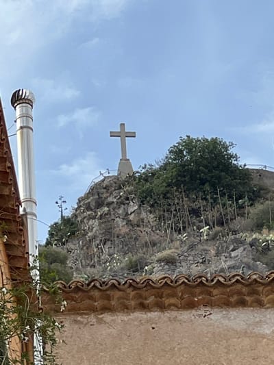 This is a photograph taken in Taormina, Italy, capturing a cross atop a rocky hill. In the foreground, a portion of a building with a tiled roof is visible. To the left, a tall, cylindrical metal structure with a cap is present, possibly a chimney or vent. The hill is covered in rocks, sparse vegetation, and several thin, upright plants. A large, dark green tree sits on top of the hill, behind the cross. The sky is partly cloudy and light blue.