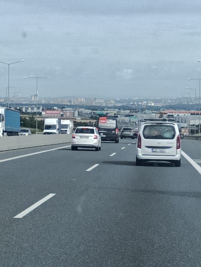 This is a photograph taken from a vehicle traveling on a multi-lane highway, likely in the vicinity of Kuří, Czechia, given the context. The sky is overcast with a light haze. The road is paved with asphalt and marked with white lines indicating lanes.
In the foreground, the road surface is visible with lane markings. Several vehicles are present:
* On the rightmost lane, a white van with a license plate reading "BMX 9942" is in motion.
* In the lane to the left of the white van, a white passenger car is moving.
* Further ahead and to the left, a black van or SUV is on the road.
* In the far left lane, a blue semi-trailer truck is traveling.
* Behind the blue semi-trailer truck, a white vehicle, possibly another truck or van, is visible.
In the middle ground, beyond the vehicles, there is a concrete barrier separating the highway from the opposing lanes of traffic. Beyond the barrier, there is a landscape with trees and buildings. A sign with "tescoma" is visible on one of the buildings. In the background, a cityscape with numerous buildings and structures can be seen, stretching towards the horizon. Streetlights are also visible along the highway. The overall scene depicts a typical highway environment with moderate traffic under a cloudy sky.