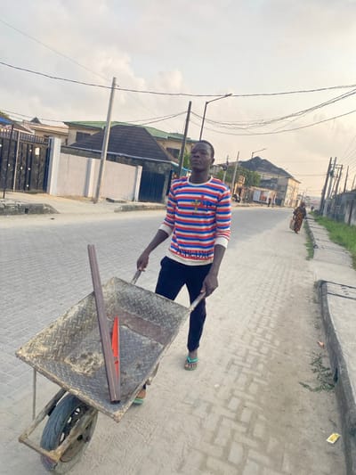 A man is pulling a metal wheelbarrow on a paved street. He wears a striped shirt, dark pants, and sandals. The wheelbarrow contains long metal rods. Buildings and utility poles line the street. A woman is walking away from the viewer in the distance. The sky is overcast.