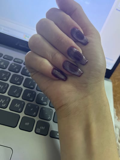 A hand with long, purple cat-eye manicured nails rests on a laptop keyboard. The hand is fair-skinned and angled towards the viewer. The nails are the focal point, showcasing a distinctive cat-eye effect. A laptop screen glows to the left. The keyboard features a mix of English and Arabic characters. The keys are dark gray. The image's perspective is from above, highlighting the nails, hand, and laptop interface.
