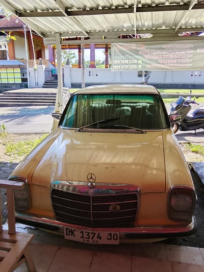 The image shows the front of a yellow Mercedes-Benz sedan parked under a metal awning. The car has a chrome grille with the Mercedes-Benz logo at the top and a license plate that reads "DK 1374 JO" and "08 30". Behind the car, a dark-colored scooter is visible. In the background, there is a building with traditional Balinese architecture, featuring intricate carvings and purple and white checkered curtains. Steps lead up to the building. A banner with text, some of which is in English and some in a language that appears to be Indonesian, hangs from the awning. The scene appears to be in Tabanan, Indonesia, as indicated by the license plate format and the architectural style. The ground in front of the car is tiled, and there is some vegetation on the left side of the image. The overall impression is of a vehicle parked in a sheltered area in a location with a strong cultural identity.