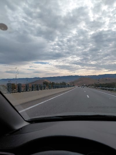 A car travels on a paved road beside a guardrail. Beyond the rail lies a mountainous landscape. A dramatic, cloudy sky covers most of the view. The car's dashboard and part of the steering wheel are visible in the foreground, suggesting the photo was taken from inside the vehicle. The road is marked with a white line along its edge.